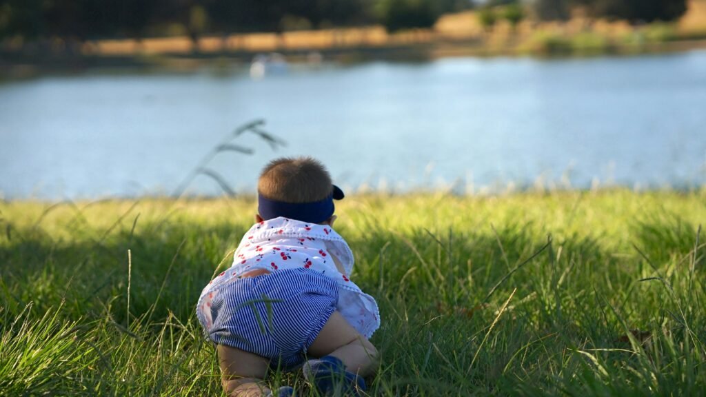 child in green and white stripe shirt sitting on green grass field near body of water child in green and white stripe shirt sitting on green grass field near body of water
